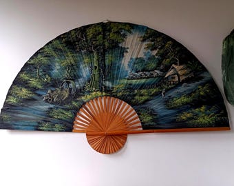 May include: A large, semi-circular wall fan featuring a painted landscape scene with a thatched-roof hut, lush green trees, and two oxen pulling a cart. The fan has a light brown wooden frame with radiating slats at the base and a dark fabric canopy.
