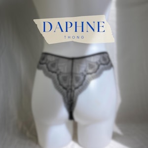 May include: A black lace thong featuring a scalloped edge design. A beige rectangular label is positioned above the garment with the blue text DAPHNE THONG printed on it. The thong is displayed on a white mannequin torso.
