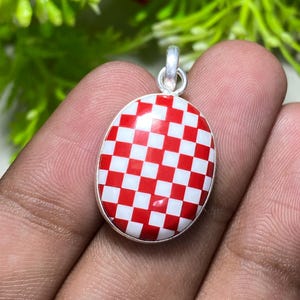 May include: Oval pendant with a red and white checkered pattern set in a silver-colored metal. The pendant has a small loop at the top for a chain. The background is blurred with green and red foliage.