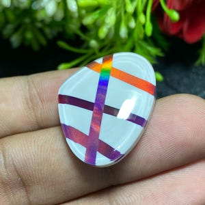 May include: A white, oval-shaped cabochon with a colorful, intersecting design. The design features a rainbow-colored stripe and intersecting red and purple stripes. The cabochon is smooth and polished, reflecting light.