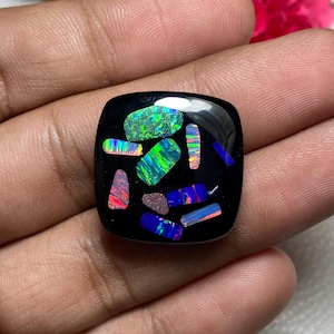 May include: A square, black gemstone with a glossy finish. The stone is inlaid with colorful, iridescent opal fragments in shades of green, blue, orange, and pink. The opal pieces are arranged in a mosaic pattern.