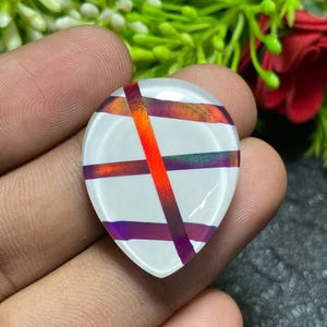 May include: A teardrop-shaped, white cabochon with intersecting bands of red, orange, and purple. The bands create a striped pattern across the surface. The cabochon is held in a hand, with a blurred green and red background.