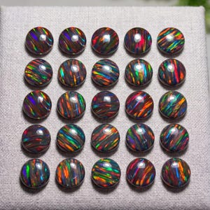 May include: A collection of twenty-five round, dark-toned cabochon gemstones arranged in a five-by-five grid. Each stone features a smooth, polished surface with vibrant, multi-colored iridescent flashes of red, green, and blue light.