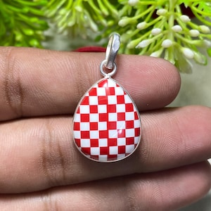 May include: A teardrop-shaped pendant with a red and white checkered pattern. The pendant has a silver-colored bail for attaching to a chain. The background is blurred, with green and white foliage.