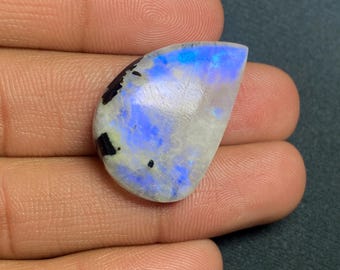 Könnte beinhalten: Ein tropfenförmiger, irisierender Mondstein-Cabochon mit blauem und weißem Schimmer. Der Stein hat schwarze Einschlüsse und ist in einer polierten, glatten, ovalen Form gefasst.