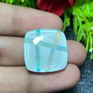 May include: A square, light blue gemstone with iridescent stripes in a grid pattern. The gemstone is held in a hand, with a blurred background of green and red foliage.