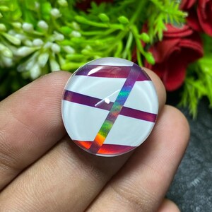 May include: A round, white-based cabochon with a crisscross pattern of purple and iridescent stripes. The iridescent stripes reflect a rainbow of colors. The cabochon is held in a hand, with green and red floral elements in the background.