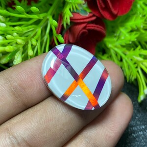 May include: An oval-shaped, white cabochon with a crisscross pattern of orange and purple stripes. The cabochon is smooth and reflective, with the stripes appearing to overlap. The background features green foliage and red flowers.