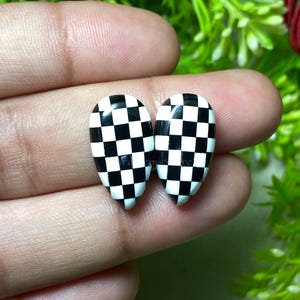 May include: Two teardrop-shaped nail art designs with a black and white checkered pattern. The nail art is held between fingers, showcasing the glossy finish and the contrasting colors. The design is a classic checkerboard.