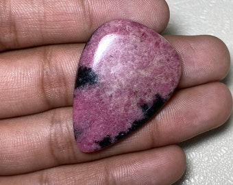 Könnte beinhalten: Ein rosa-schwarzer Cabochon-Edelstein in Tropfenform. Der Stein hat eine glatte, polierte Oberfläche und ist in einen silbernen Ring gefasst.