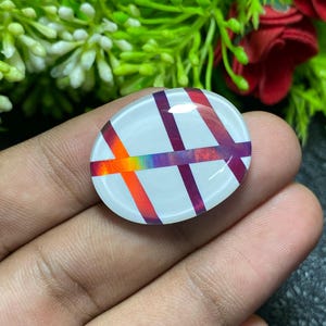 May include: An oval-shaped, white cabochon with a colorful, crisscrossing design. The design features bands of orange, red, purple, and rainbow hues. The cabochon is held in a hand, with a blurred background of greenery and red flowers.