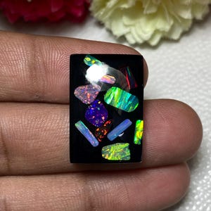 Może przedstawiać: Prostokątny czarny wisiorek z kamienia szlachetnego inkrustowany iryzującymi fragmentami opalu. Opale prezentują gamę kolorów, w tym niebieski, zielony, fioletowy i pomarańczowy. Wisiorek trzymany jest między palcami.