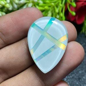 May include: A teardrop-shaped, translucent gemstone with iridescent, intersecting bands of blue, green, and yellow. The stone is held in a hand, with a blurred background of green foliage and a red flower.