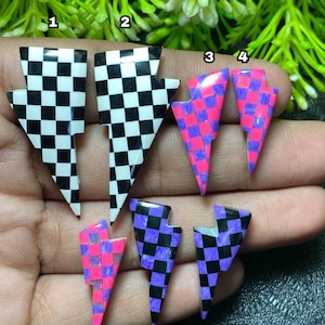Può includere: Una collezione di sette orecchini a forma di fulmine. Gli orecchini presentano motivi a scacchi in bianco e nero, rosa e viola e viola e nero. Gli orecchini sono esposti su uno sfondo verde e scuro.