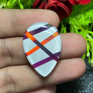 May include: A teardrop-shaped, white gemstone with intersecting bands of orange, purple, and dark purple. The gemstone is held in a hand, with green foliage and a red flower in the background.