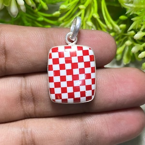 May include: A square pendant with a red and white checkered pattern. The pendant has a silver-colored bail for attaching to a chain. The background features green foliage.