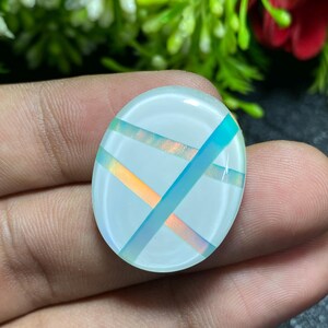 May include: Oval-shaped, iridescent gemstone with a white base and intersecting bands of blue, orange, and purple. The gemstone is held in a hand, with green foliage and a red flower in the background.