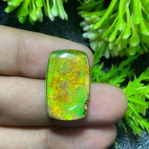 May include: A rectangular gemstone with a vibrant display of colors, including gold, green, and hints of orange. The stone is held between fingers, with green foliage in the background. The gemstone has a polished, reflective surface.