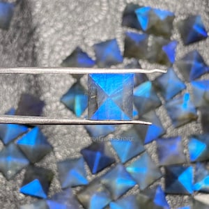Puede incluir: Primer plano de una piedra preciosa de labradorita azul, cortada en forma de pirámide cuadrada. La piedra preciosa está siendo sostenida por unas pinzas y está rodeada de otras piedras preciosas similares.
