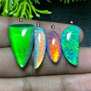 Può includere: Quattro pietre preziose di opale lucide in varie forme e colori. Le pietre preziose mostrano tonalità vibranti di verde, blu, arancione e giallo. Gli opali sono tenuti in una mano, mostrando i loro motivi unici e le qualità iridescenti.