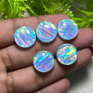 Puede incluir: Cinco ópalos redondos y pulidos. Cada ópalo tiene un color base azul claro con destellos iridiscentes de rosa, verde y amarillo. Los ópalos se muestran contra una mano, con follaje verde en el fondo.