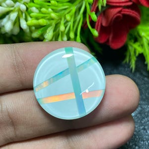 May include: A round, light blue gemstone with intersecting bands of orange, teal, and blue. The gemstone is held in a hand, with green foliage and red flowers in the background. The gemstone has a polished, reflective surface.