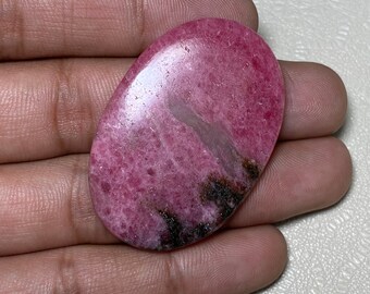 May include: A pink and black oval-shaped cabochon gemstone with a polished surface. The stone has a swirling pattern of pink and black.