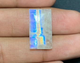 May include: A rectangular, iridescent gemstone with a white base and blue and gold flashes of color. The gemstone is approximately 1 inch long and 1/2 inch wide.