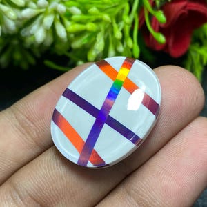 May include: Oval-shaped white gemstone with a crisscross pattern of orange, purple, and iridescent rainbow stripes. The gemstone is held in a hand, with green foliage and a red flower in the background.