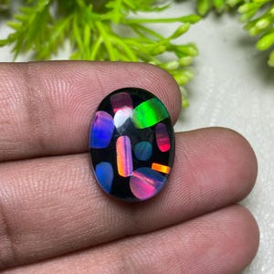 Könnte beinhalten: Ein ovaler schwarzer Opal-Edelstein mit einer lebendigen Farbgebung. Der Opal zeigt Blitze in Blau, Grün, Rot und Orange. Der Edelstein wird in einer Hand gehalten, was seine polierte Oberfläche und sein einzigartiges Farbspiel zeigt.