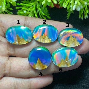 May include: Five oval and circular cabochons featuring a blue iridescent background with a gold-colored mountain range graphic at the base. Each piece is numbered one through five in black text, displaying a smooth, polished surface finish.