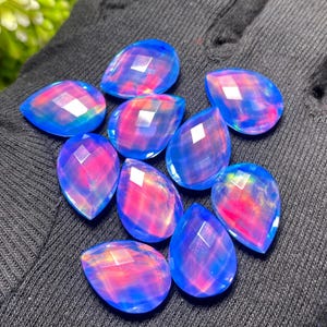 May include: A collection of ten faceted, teardrop-shaped gemstones. The gems have a vibrant blue hue with internal flashes of pink and red. The stones are arranged on a dark, textured surface.
