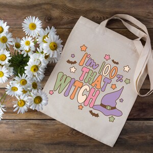 May include: A white canvas tote bag with a purple witch hat and the text "I'm 100% That Witch" in colorful letters. The bag has a brown wooden background with white daisies.