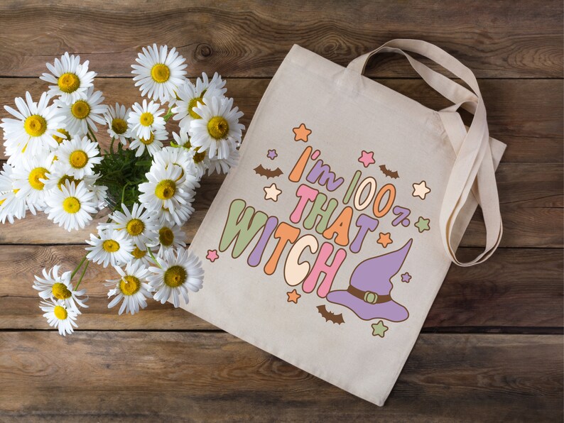 May include: A white canvas tote bag with a purple witch hat and the text "I'm 100% That Witch" in colorful letters. The bag has a brown wooden background with white daisies.