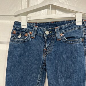 May include: Blue denim jeans with orange stitching and metal button and rivets. The jeans are hanging on a white hanger. The jeans have front pockets and belt loops.