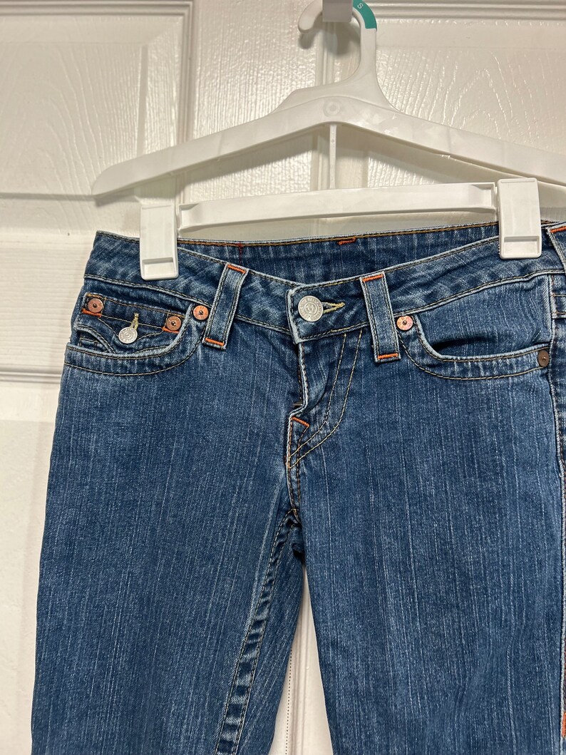 May include: Blue denim jeans with orange stitching and metal button and rivets. The jeans are hanging on a white hanger. The jeans have front pockets and belt loops.