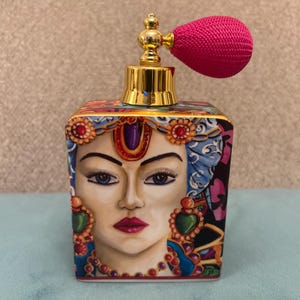 Könnte beinhalten: Dekorative Parfümflasche mit goldfarbenem Deckel und rosa Zerstäuber. Die Flasche zeigt ein gemaltes Gesicht einer Frau mit blauen Augen, roten Lippen und aufwendigem Schmuck. Die Flasche ist quadratisch und hat bunte Blumenmuster.
