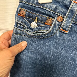 May include: Close-up of blue denim jeans with orange stitching and metal rivets. The jeans feature a small front pocket with a silver button and a decorative design. The fabric has a worn, faded appearance.