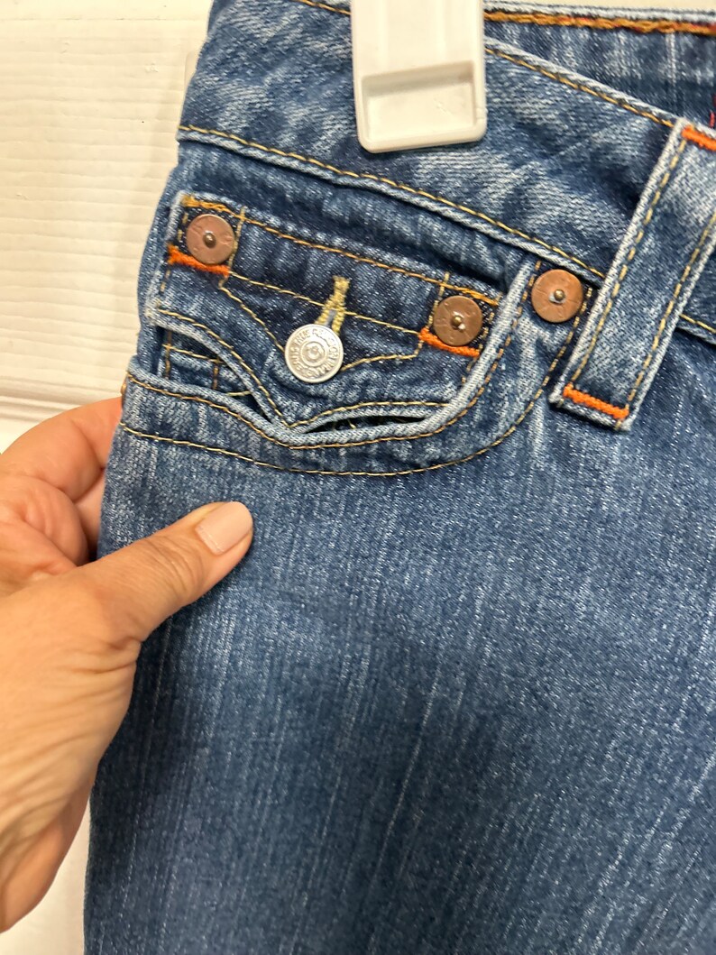 May include: Close-up of blue denim jeans with orange stitching and metal rivets. The jeans feature a small front pocket with a silver button and a decorative design. The fabric has a worn, faded appearance.