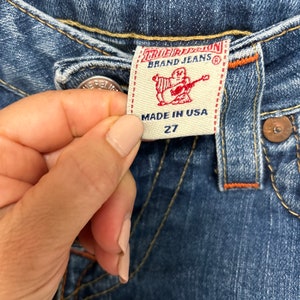 May include: Close-up of blue denim jeans with a True Religion Brand Jeans label. The label is cream-coloured with red text and a graphic of a figure playing a guitar. The jeans are size 27 and made in the USA.