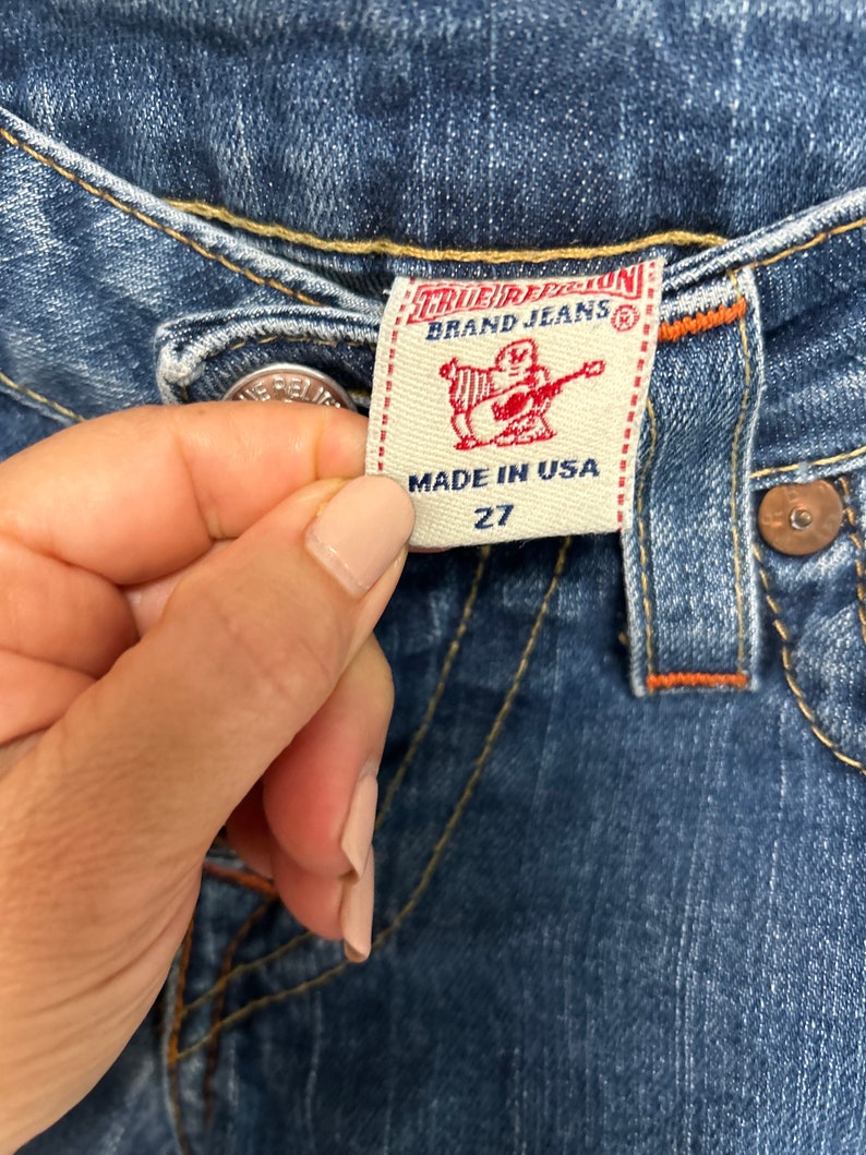 May include: Close-up of blue denim jeans with a True Religion Brand Jeans label. The label is cream-coloured with red text and a graphic of a figure playing a guitar. The jeans are size 27 and made in the USA.