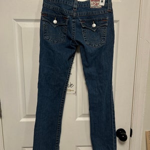 May include: Dark wash denim jeans with a classic five-pocket design. Features decorative stitching, silver button closures, and a brand label on the back. The jeans are hanging on a white hanger.