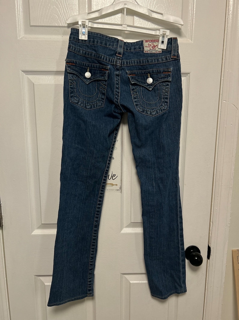 May include: Dark wash denim jeans with a classic five-pocket design. Features decorative stitching, silver button closures, and a brand label on the back. The jeans are hanging on a white hanger.