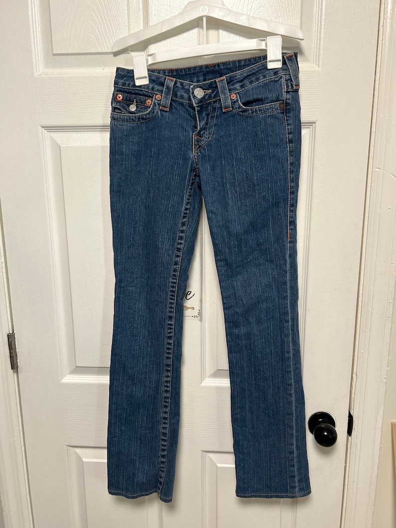May include: Blue denim jeans with a classic five-pocket design, featuring a button and zip closure. The jeans are hanging on a white hanger. The stitching is a contrasting orange colour.