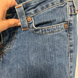 May include: Close-up of blue denim jeans with orange stitching. The jeans feature a classic five-pocket design, with a small pocket on the front and two pockets on the back. The jeans are hanging on a white hanger.