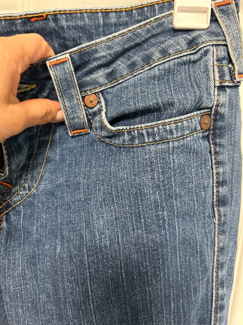 May include: Close-up of blue denim jeans with orange stitching. The jeans feature a classic five-pocket design, with a small pocket on the front and two pockets on the back. The jeans are hanging on a white hanger.