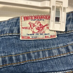 May include: Close-up of blue denim jeans with orange and yellow stitching. The True Religion brand label is visible, featuring a graphic of a Buddha playing a guitar and the text "Fashion for the Senses."