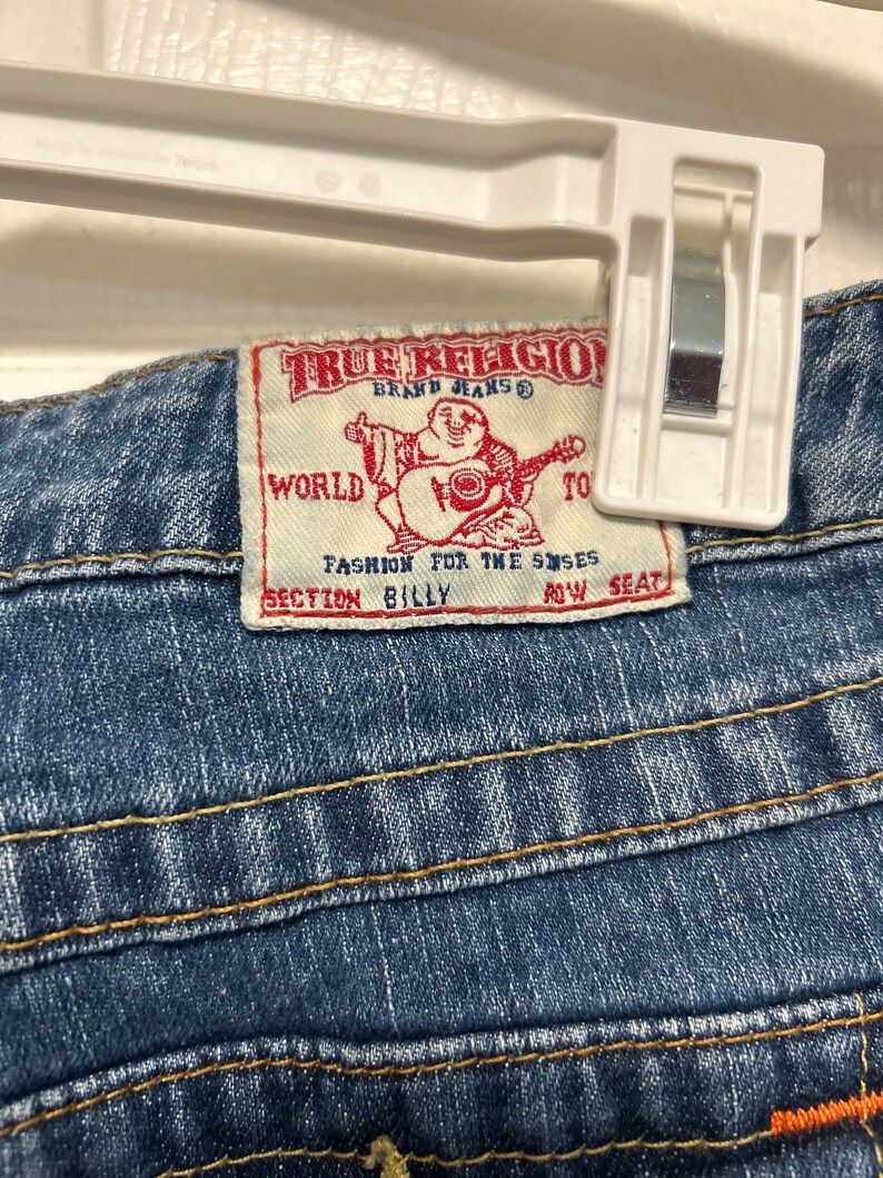 May include: Close-up of blue denim jeans with orange and yellow stitching. The True Religion brand label is visible, featuring a graphic of a Buddha playing a guitar and the text "Fashion for the Senses."
