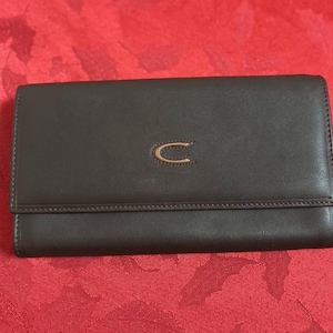 May include: A brown leather wallet with a gold-colored logo in the center. The wallet has a rectangular shape with a flap closure. The stitching is visible around the edges. The wallet is on a red surface.