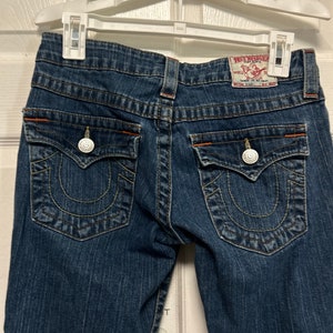 May include: Dark blue denim jeans with decorative stitching and orange accents. The jeans have two back pockets with silver button closures and a True Religion brand label.